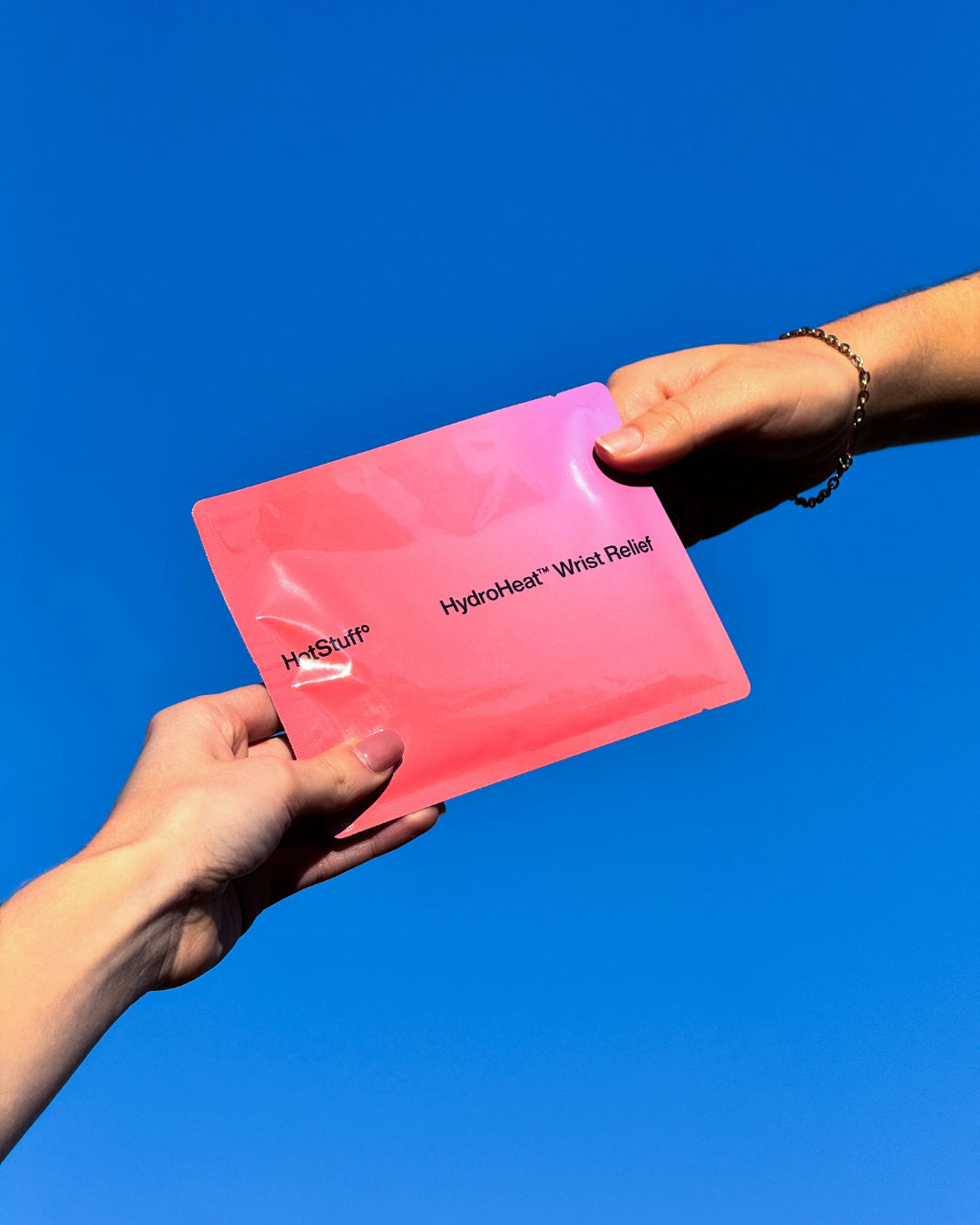 Pink Hotstuff HydroHeat Wrist Relief patch held by two hands against a blue sky, highlighting portable wrist pain relief.